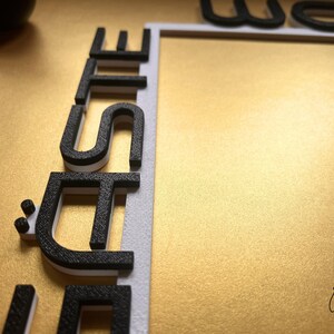 May include: Close-up of a gold surface with the word "SITE" in black and white 3D letters. The letters are positioned vertically along a white border. The word "WE" is partially visible at the top. The image has a textured gold background.