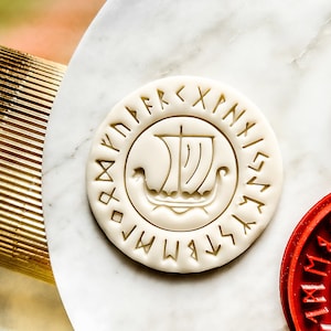 May include: White cookie cutter with a Viking ship design surrounded by runes. The cookie cutter is on a white marble surface.