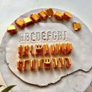 May include: A set of orange plastic alphabet cookie cutters arranged in a semi-circle above and below a white clay slab. The clay slab features the embossed letters A through I in a gothic font style, with additional loose cutters scattered around.