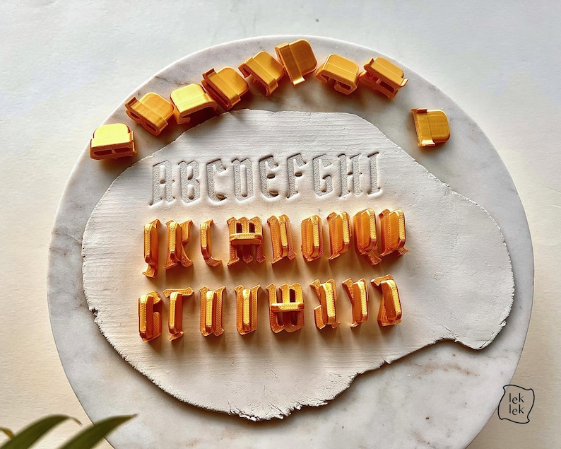 May include: A set of orange plastic alphabet cookie cutters arranged in a semi-circle above and below a white clay slab. The clay slab features the embossed letters A through I in a gothic font style, with additional loose cutters scattered around.