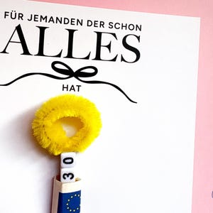 May include: A white card with the black text F&Uuml;R JEMANDEN DER SCHON ALLES HAT and a black bow graphic. Below the text is a yellow fuzzy pipe cleaner circle, two white square beads with the number 30, and a blue and white paper roll.