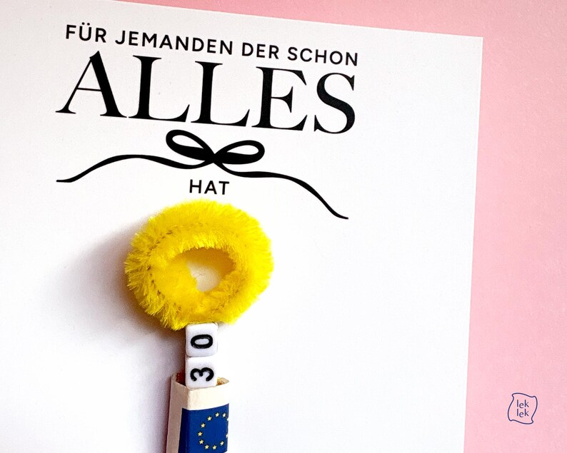 May include: A white card with the black text F&Uuml;R JEMANDEN DER SCHON ALLES HAT and a black bow graphic. Below the text is a yellow fuzzy pipe cleaner circle, two white square beads with the number 30, and a blue and white paper roll.
