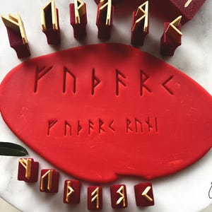 Puede incluir: Un conjunto de herramientas de sello rúnico rojas y doradas dispuestas alrededor de una pieza de masa roja circular, plana y grande. Los sellos presentan varios símbolos Elder Futhark, que también están impresos en la superficie de la masa en dos filas.
