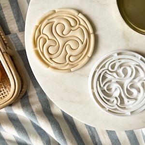 May include: Two white cookie cutters with a Celtic knot design. The cookie cutters are on a white marble surface.
