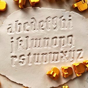 May include: A set of yellow plastic alphabet stamps used to press lowercase letters into a flat, off-white clay surface. The letters a through z are clearly imprinted in the clay, with several individual yellow stamp tools scattered around the edges.