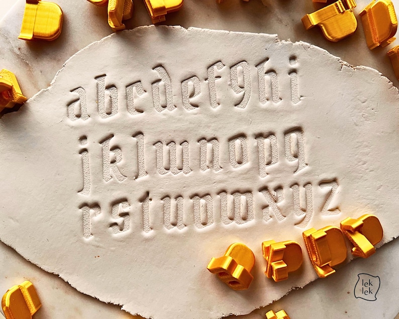 May include: A set of yellow plastic alphabet stamps used to press lowercase letters into a flat, off-white clay surface. The letters a through z are clearly imprinted in the clay, with several individual yellow stamp tools scattered around the edges.