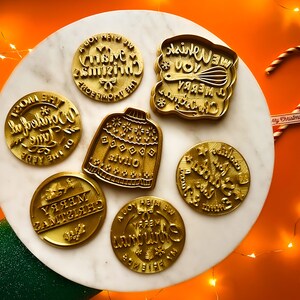 May include: Set of 7 gold-colored cookie cutters with Christmas-themed designs. The designs include "We Wish You A Merry Christmas", "Oh The Wonderful Time Of The Year", "Merry Christmas", "It's The Most Wonderful Time Of The Year", "Jingle All The Way", "Have Yourself A Merry Little Christmas", and a sweater.
