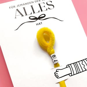 May include: A white card with the German text "F&Uuml;R JEMANDEN DER SCHON ALLES HAT" and the word "HAT" below a black bow. A yellow pipe cleaner with the number "70" on it is attached to the card, with a drawing of a hand.