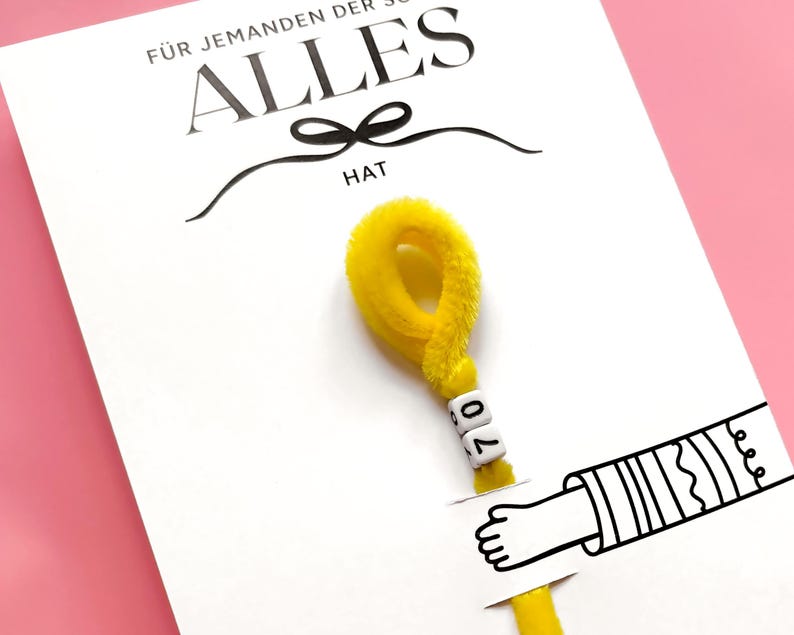 May include: A white card with the German text "F&Uuml;R JEMANDEN DER SCHON ALLES HAT" and the word "HAT" below a black bow. A yellow pipe cleaner with the number "70" on it is attached to the card, with a drawing of a hand.