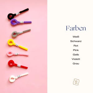Op de afbeelding: Zeven pluizige pijpenragers in zwart, paars, grijs, geel, roze, rood en wit, elk met een lusvormig uiteinde en een klein wit label met een nummer. De tekst "Farben" staat boven een lijst met kleuren: Wei&szlig;, Schwarz, Rot, Pink, Gelb, Violett, Grau.
