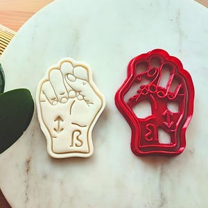 May include: A beige cookie cutter in the shape of a fist with the ASL sign for the letter "B" and an arrow symbol. Next to it is a red plastic cookie cutter in the same shape. The items are on a marble surface.