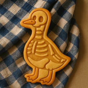 May include: A golden-brown cookie shaped like a duck, featuring a detailed skeleton design. The cookie rests on a blue and white checkered cloth, creating a contrast of textures and colors. The duck's skeleton is intricately outlined.