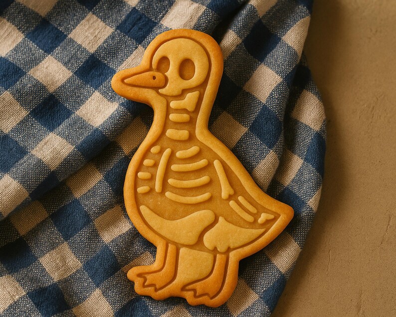 May include: A golden-brown cookie shaped like a duck, featuring a detailed skeleton design. The cookie rests on a blue and white checkered cloth, creating a contrast of textures and colors. The duck's skeleton is intricately outlined.