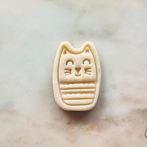 May include: A beige cat-shaped cookie cutter with a smiling face and striped body. The cookie cutter is made of plastic and has the text "lek lek" in the bottom right corner.
