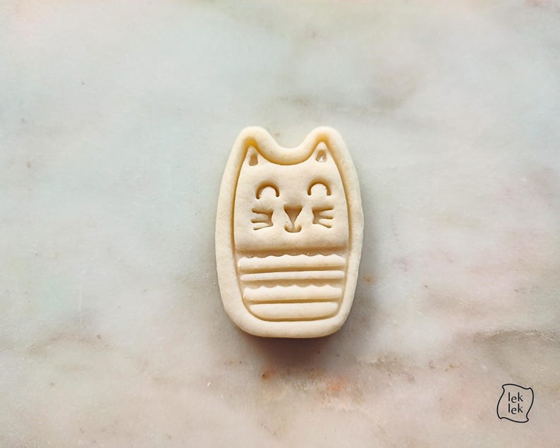 May include: A beige cat-shaped cookie cutter with a smiling face and striped body. The cookie cutter is made of plastic and has the text "lek lek" in the bottom right corner.