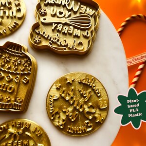 May include: A set of five gold-colored cookie cutters with Christmas-themed designs. The designs include a Christmas sweater, a Christmas tree, a candy cane, and two different Christmas greetings. The cookie cutters are made of plant-based PLA plastic.