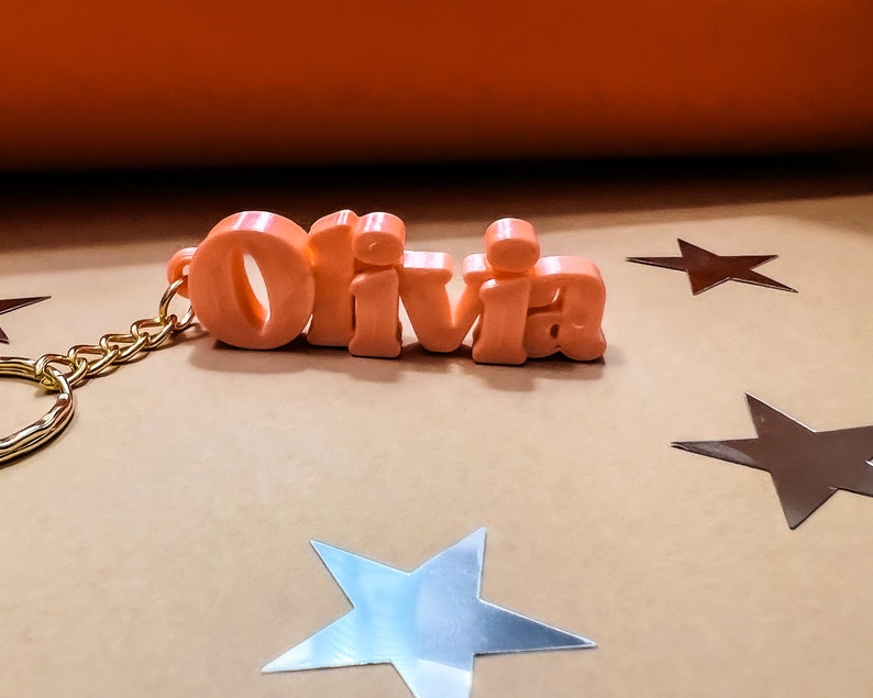 May include: A pink plastic keychain with the name "Olivia" spelled out in large letters. The keychain is attached to a gold chain.