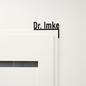 May include: Black metal door sign with the text "Dr. Imke" in a modern font. The sign is mounted on the corner of a white door and wall, creating a minimalist aesthetic. The sign is designed to be mounted on a door.