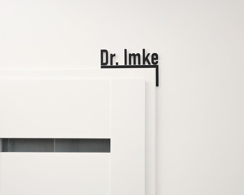May include: Black metal door sign with the text "Dr. Imke" in a modern font. The sign is mounted on the corner of a white door and wall, creating a minimalist aesthetic. The sign is designed to be mounted on a door.