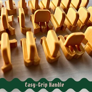 May include: A collection of gold-colored, 3D-printed plastic components, each featuring a vertical tab and a base, arranged in rows. The text "Easy-Grip Handle" is displayed in a dark green, wavy-edged banner at the bottom of the image.