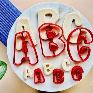 Könnte beinhalten: Rote Kunststoff-Ausstecher in Form von Buchstaben des Alphabets auf einem weißen Teller mit weißen Fondant-Buchstaben. Die Ausstecher haben die Form der Buchstaben A, B und C.
