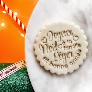 May include: A white cookie with the text "Joyeux Noel a la famille leger et bonne annee" in brown lettering. The cookie is decorated with small hearts.