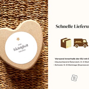 K&ouml;nnte beinhalten: Eine herzf&ouml;rmige, hellbraune Schachtel mit einem wei&szlig;en, kreisf&ouml;rmigen Etikett. Das Etikett tr&auml;gt den Text "EINE Kleinigkeit F&Uuml;R DICH" in Schwarz. Die Schachtel befindet sich auf einer gewebten, braunen Oberfl&auml;che.
