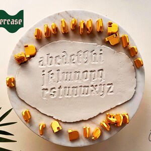 May include: A set of gold-colored lowercase letter stamps arranged around a central piece of white clay. The clay features the full alphabet from a to z pressed into its surface in a gothic font. The word lowercase is printed in white on a green banner.