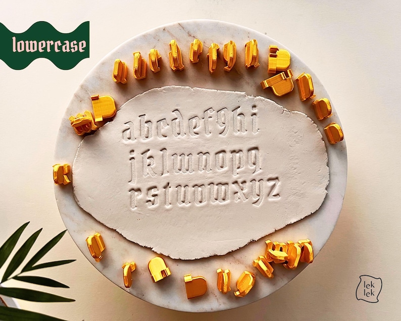 May include: A set of gold-colored lowercase letter stamps arranged around a central piece of white clay. The clay features the full alphabet from a to z pressed into its surface in a gothic font. The word lowercase is printed in white on a green banner.
