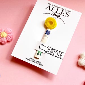 May include: A white card featuring the German text F&Uuml;R JEMANDEN DER SCHON ALLES HAT and BAUCHNABEL REINIGUNGSB&Uuml;RSTE. A small yellow pipe cleaner brush is attached to the card, held by a printed illustration of an arm. Two pink yarn flowers are present.