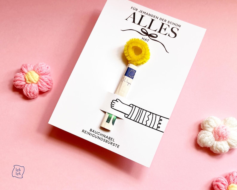 May include: A white card featuring the German text F&Uuml;R JEMANDEN DER SCHON ALLES HAT and BAUCHNABEL REINIGUNGSB&Uuml;RSTE. A small yellow pipe cleaner brush is attached to the card, held by a printed illustration of an arm. Two pink yarn flowers are present.