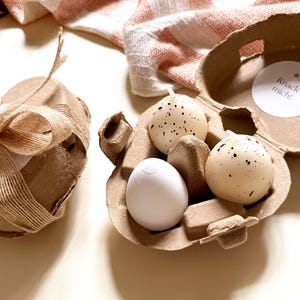Pu&ograve; includere: Una confezione regalo decorativa legata con spago e un fiocco, accanto a un cartone per uova contenente tre uova macchiate e un uovo bianco. Il cartone per uova ha un adesivo che dice "Knack mich!"