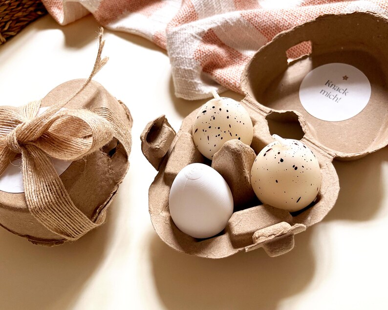 Pu&ograve; includere: Una confezione regalo decorativa legata con spago e un fiocco, accanto a un cartone per uova contenente tre uova macchiate e un uovo bianco. Il cartone per uova ha un adesivo che dice "Knack mich!"