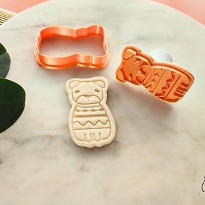 Puede incluir: Juego de cortadores de galletas de pl&aacute;stico naranja con forma de perro. El juego incluye un cortador, un sello y una galleta. La galleta tiene un dise&ntilde;o detallado de las caracter&iacute;sticas y la ropa del perro. El juego est&aacute; sobre una superficie de m&aacute;rmol.