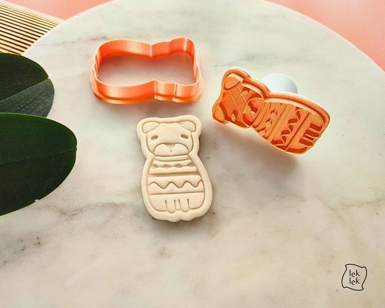 Puede incluir: Juego de cortadores de galletas de pl&aacute;stico naranja con forma de perro. El juego incluye un cortador, un sello y una galleta. La galleta tiene un dise&ntilde;o detallado de las caracter&iacute;sticas y la ropa del perro. El juego est&aacute; sobre una superficie de m&aacute;rmol.
