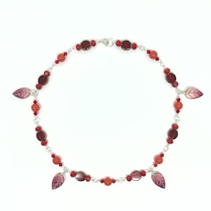 Pode incluir: Um colar com corrente prateada, contas vermelhas e pingentes de folhas. O colar apresenta uma série de contas vermelhas e contas maiores, de cor vermelho escuro e formato oval. Três pingentes em forma de folha em rosa e roxo pendem da corrente.