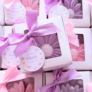 May include: White boxes with a clear window, each tied with a pink or purple satin bow. Inside each box is a flower-shaped soap in shades of pink or purple. Each box has a circular tag with text.