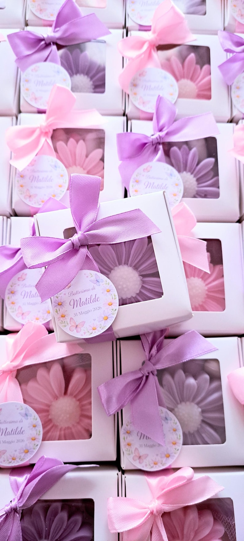 May include: White boxes with a clear window, each tied with a pink or purple satin bow. Inside each box is a flower-shaped soap in shades of pink or purple. Each box has a circular tag with text.