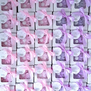 May include: Arrangement of gift boxes, each containing a flower-shaped item. Boxes are white with a clear window, tied with pink or purple satin ribbons. Each box has a circular tag with an elephant illustration.