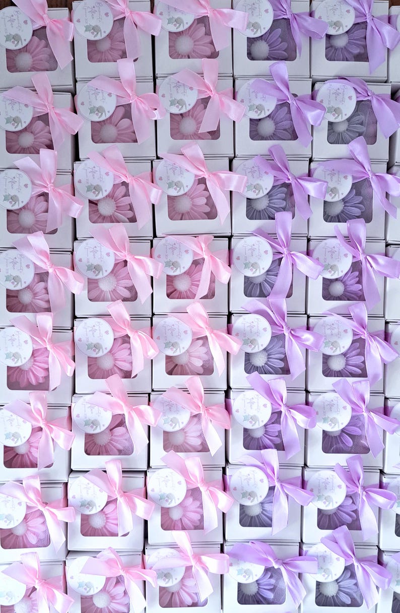 May include: Arrangement of gift boxes, each containing a flower-shaped item. Boxes are white with a clear window, tied with pink or purple satin ribbons. Each box has a circular tag with an elephant illustration.