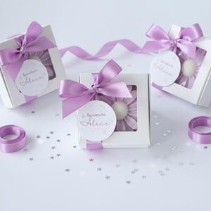 May include: White gift boxes with a window, each tied with a lavender satin bow. Inside each box is a lavender flower. Each box has a round tag that says "Benvenuta Alice". Lavender ribbon and star-shaped confetti complete the presentation.