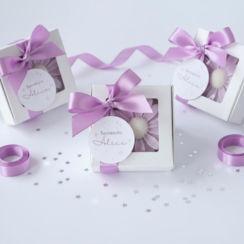 May include: White gift boxes with a window, each tied with a lavender satin bow. Inside each box is a lavender flower. Each box has a round tag that says "Benvenuta Alice". Lavender ribbon and star-shaped confetti complete the presentation.
