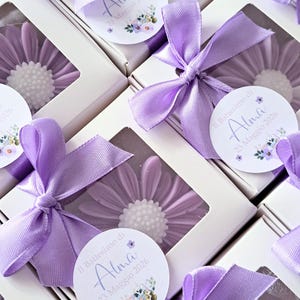 May include: Daisy-shaped soaps in white boxes, each adorned with a lavender ribbon bow. The soaps are lavender with a white center. Each box has a label that reads "Il Battesimo di Alma 23 Maggio 2026."