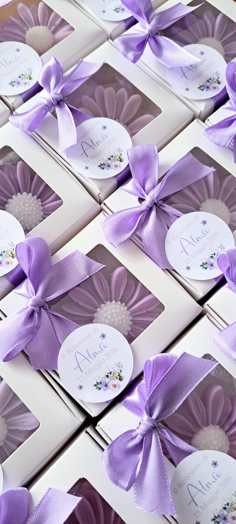 May include: Daisy-shaped soaps in white boxes, each adorned with a lavender ribbon bow. The soaps are lavender with a white center. Each box has a label that reads "Il Battesimo di Alma 23 Maggio 2026."