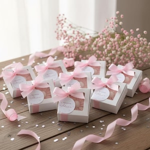 May include: White gift boxes with a clear window, tied with pink satin bows. Each box has a round tag that says "Bienvenida Alice". Pink ribbon and flowers are scattered around the boxes on a wooden surface.