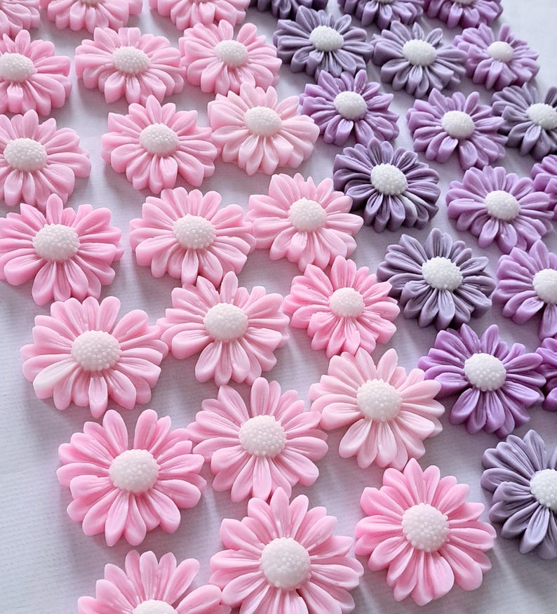 May include: An assortment of daisy-shaped embellishments in various shades of pink and purple. Each flower has a white center and detailed petals. These decorative elements are ideal for crafts and DIY projects.