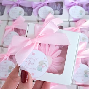 Puede incluir: Cajas de regalo blancas atadas con lazos de satén rosa, cada una con un objeto en forma de margarita rosa. Una etiqueta redonda con una ilustración de conejito y texto está adjunta. Las cajas están dispuestas en filas.
