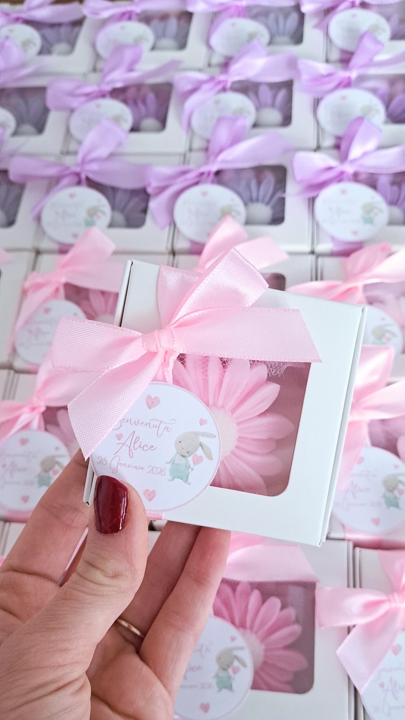 May include: White gift boxes tied with pink satin bows, each containing a pink daisy-shaped item. A round tag with a bunny illustration and text is attached to each box. The boxes are arranged in rows.