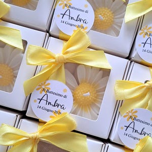 May include: White daisy-shaped candles in clear boxes, tied with yellow ribbons. Each box has a label with the text "Il Battesimo di Ambra 14 Giugno 2026". The candles are a charming gift.