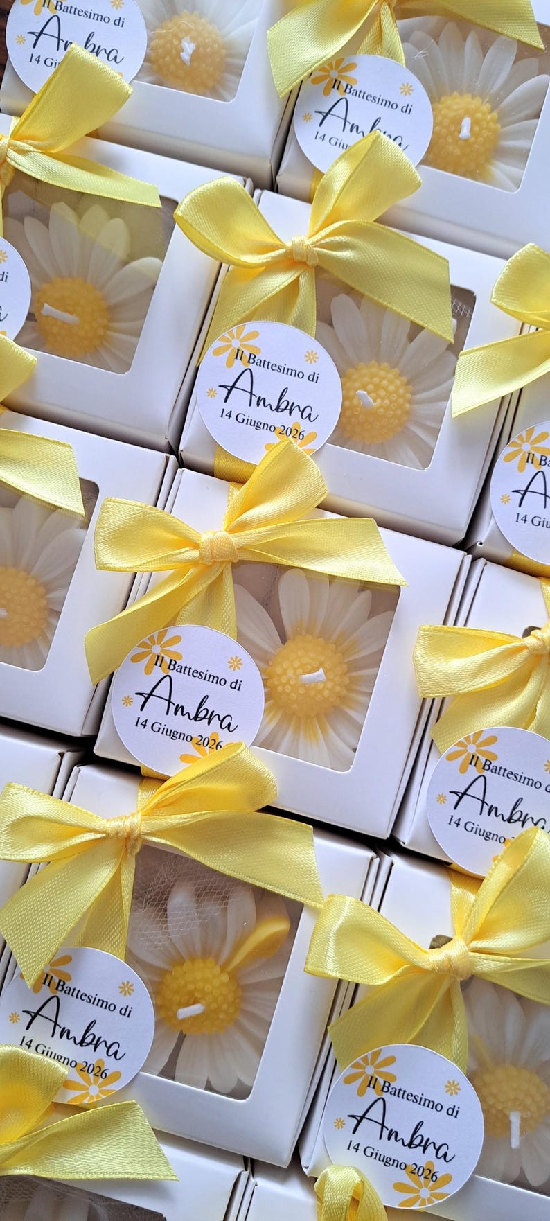 May include: White daisy-shaped candles in clear boxes, tied with yellow ribbons. Each box has a label with the text "Il Battesimo di Ambra 14 Giugno 2026". The candles are a charming gift.